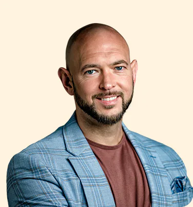 Smiling bald man with blue eyes and a short beard wearing a light blue plaid blazer over a brown shirt, posing against a light beige background.